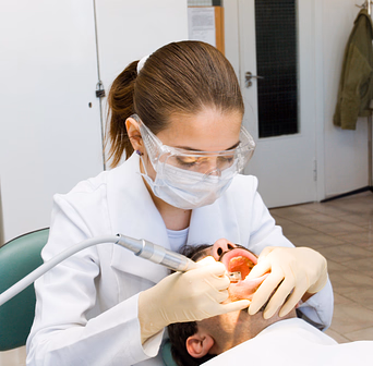 Dentist in safety glasses and mask using dental drill on patient lying back in chair.