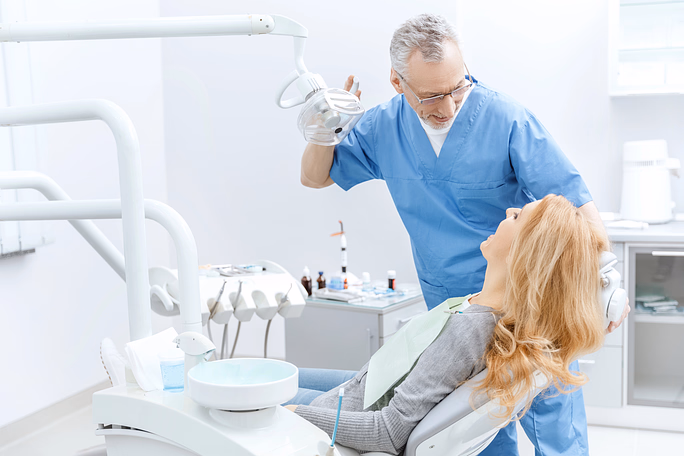 Dentist in blue scrubs adjusts overhead light while a woman reclines in the dental chair with a bib.