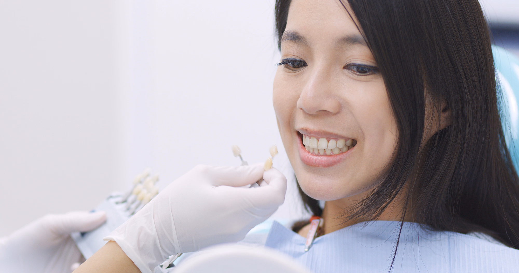 Mujer joven sonriendo ante una guía de colores dental sostenida por un dentista con guantes para igualar el color de su diente.
