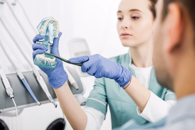 Dentista con guantes azules demostrando la técnica de cepillado en un modelo dental a un paciente en una clínica dental.