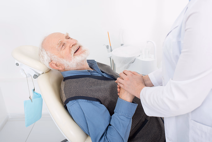 Hombre mayor sonriente recostado en una silla médica o dental, tomado de la mano con un profesional de la salud con bata blanca.
