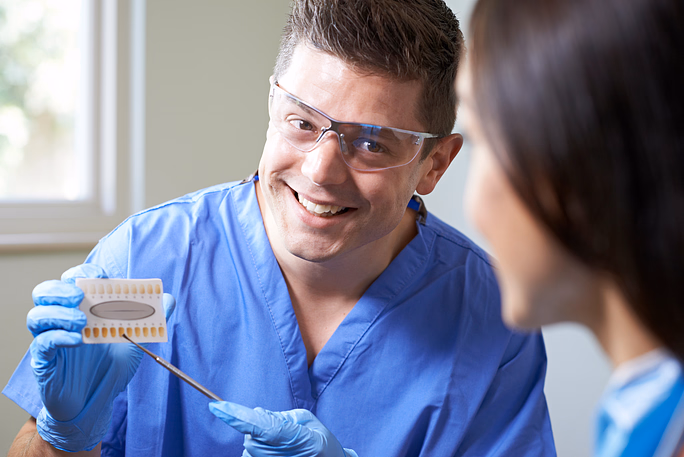 Smiling male dentist in blue scrubs and protective glasses showing a dental shade guide to a patient.