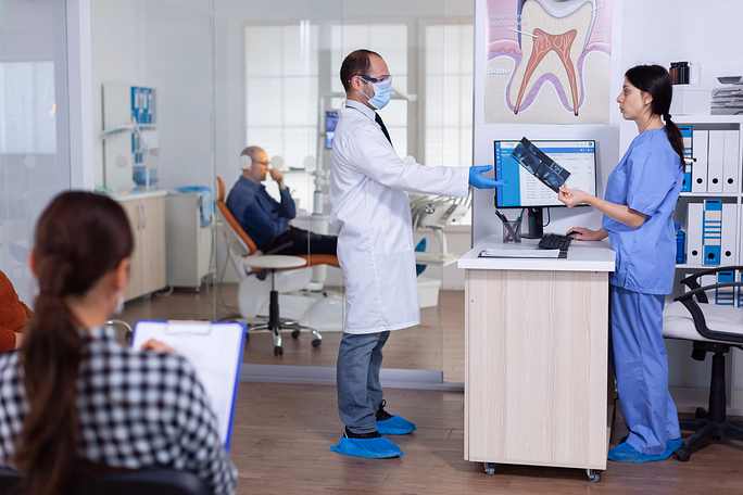 Male dentist wearing a mask and gloves discussing a dental X-ray with a female dental assistant in a clinic where a patient waits in the background.