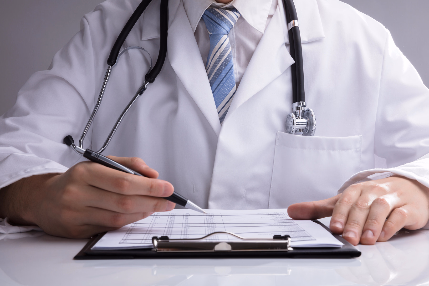 Doctor con bata blanca y corbata a rayas, sosteniendo un bolígrafo y escribiendo en un portapapeles con documentos médicos, con un estetoscopio alrededor del cuello.