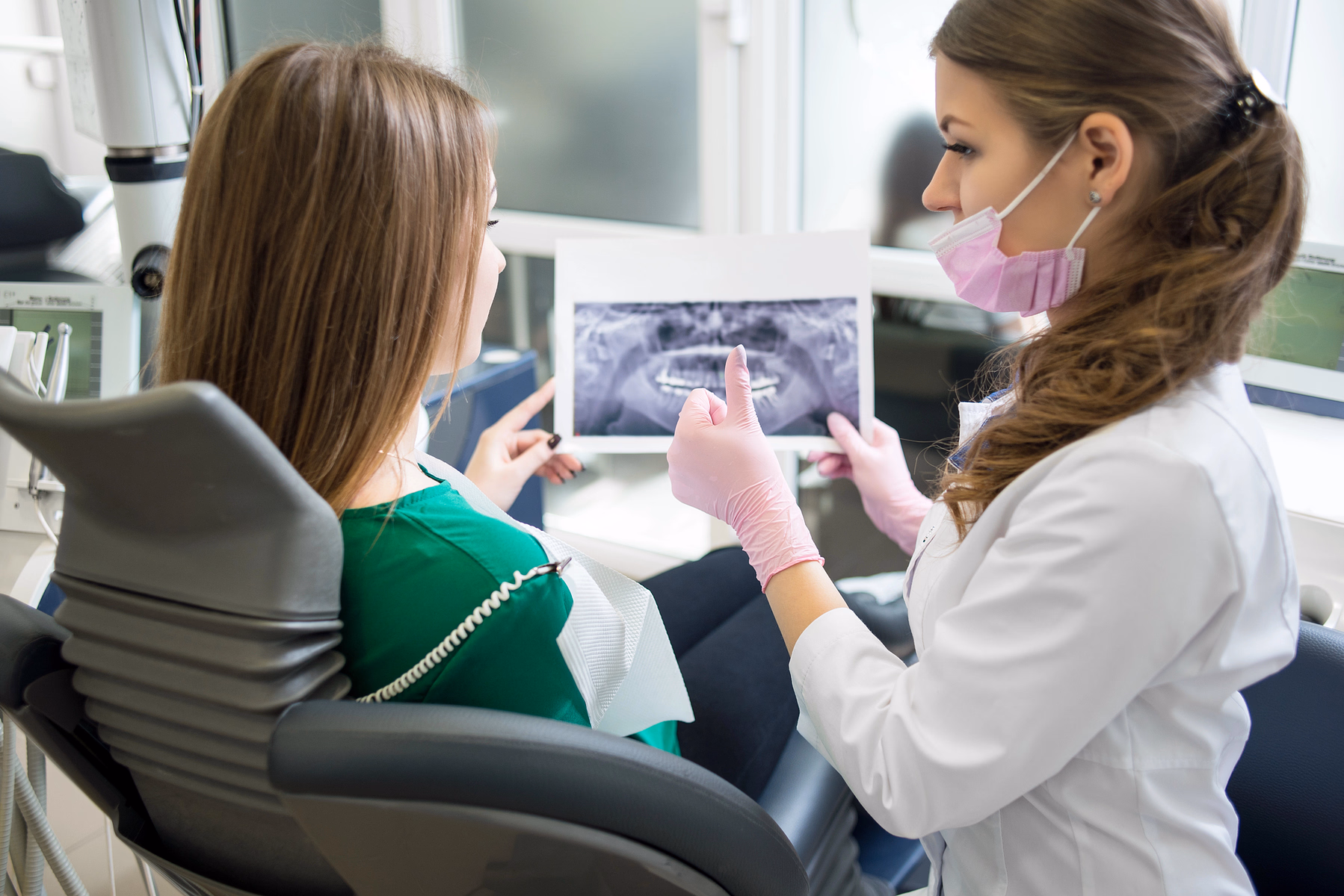 Dentista con bata blanca, guantes rosados y mascarilla mostrando una radiografía dental a una paciente sentada en una silla dental.