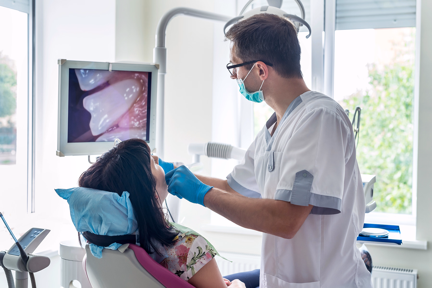 Dentista con mascarilla y guantes examinando la boca de un paciente con una cámara dental que muestra un primer plano de los dientes en un monitor.