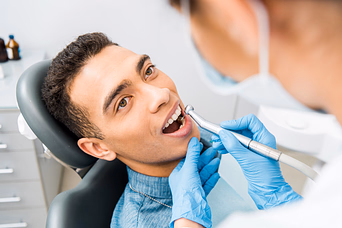 Dentist wearing blue gloves examining a smiling male patient's teeth with dental tools in a clinic.