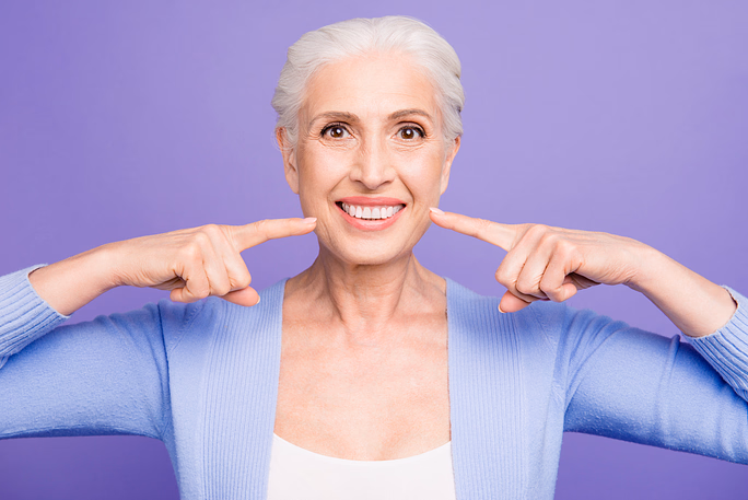 Smiling older woman with gray hair wearing a light blue cardigan pointing at her smile against a purple background.