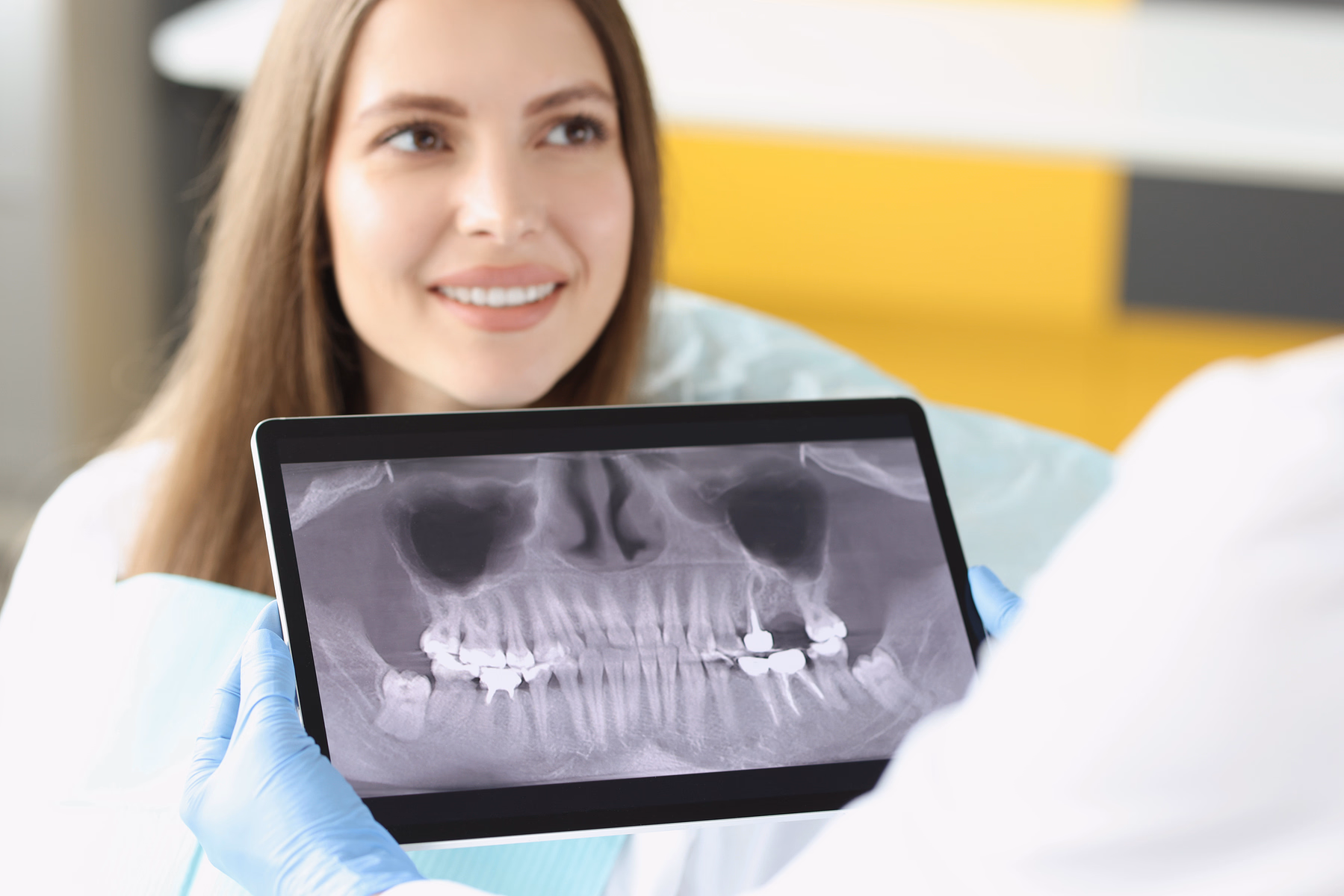 Dentist wearing blue gloves holding a tablet showing a dental X-ray near a smiling female patient.