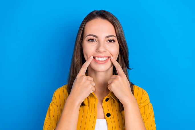 Mujer joven sonriente con camisa amarilla señalando sus dientes con ambos índices sobre un fondo azul.