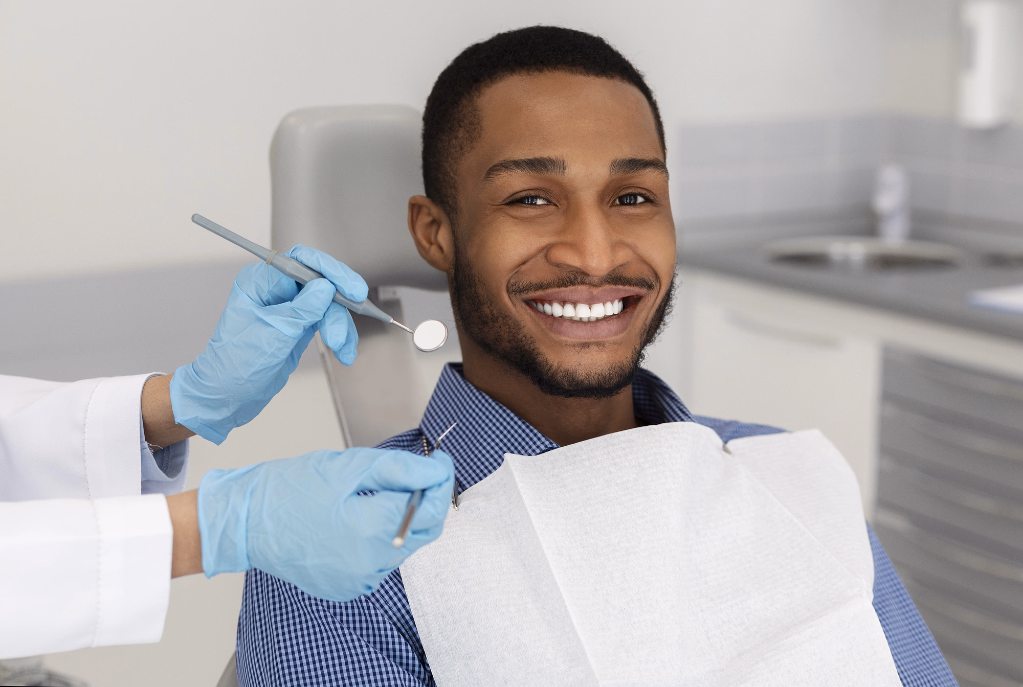 Joven sonriente sentado en una silla dental con las manos enguantadas de un dentista sosteniendo instrumentos dentales cerca de él.