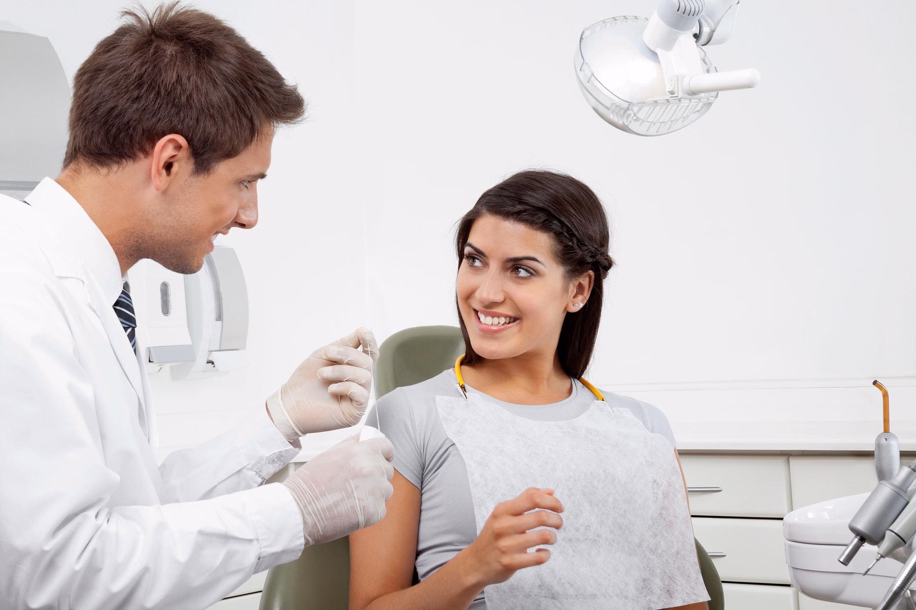 Dentista con bata blanca sosteniendo hilo dental y hablando con una paciente sonriente sentada en una silla dental.