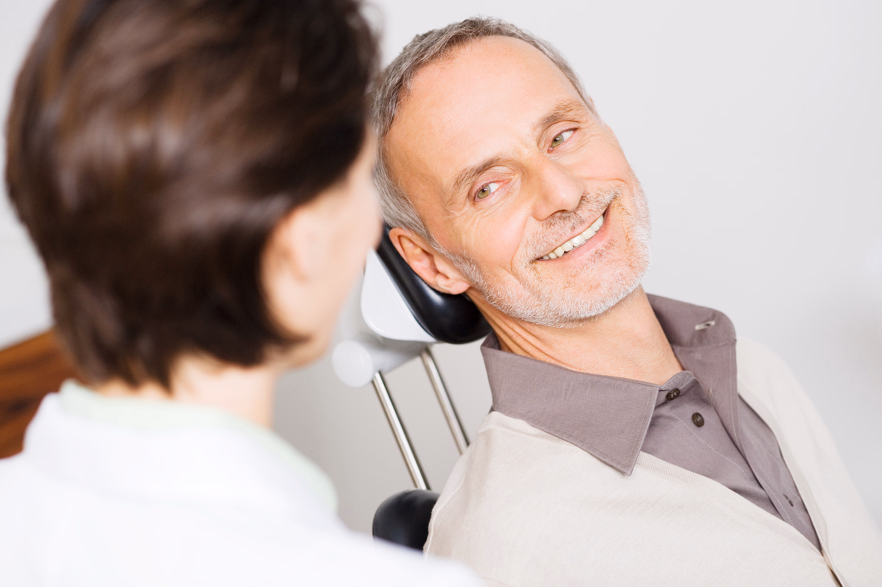 Smiling elderly man with gray hair and beard sitting in a chair, looking at a healthcare professional.