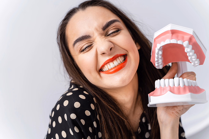 Woman with red lipstick smiling widely while holding a dental model showing gums and teeth.