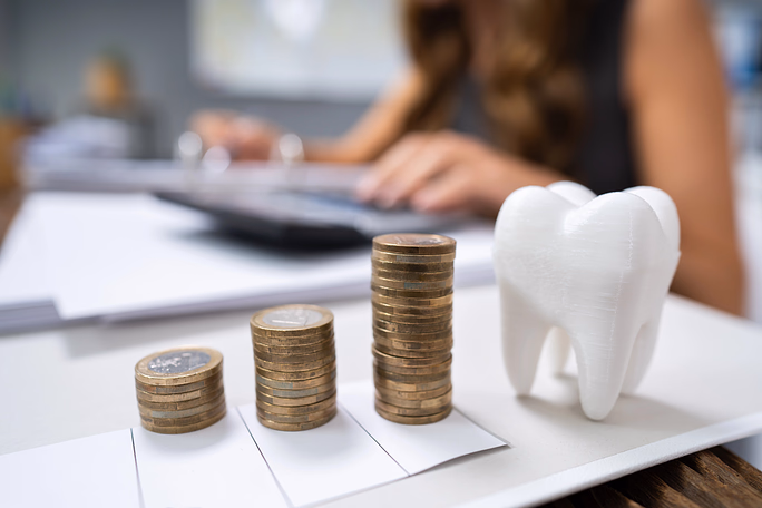 Stacks of coins increasing in height next to a white tooth model on a desk with a person using a calculator in the background.