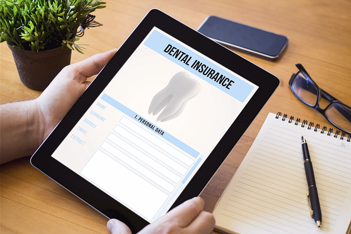 Person holding a tablet displaying a dental insurance form with fields for personal data, next to a notebook, pen, glasses, smartphone, and potted plant on a wooden table.
