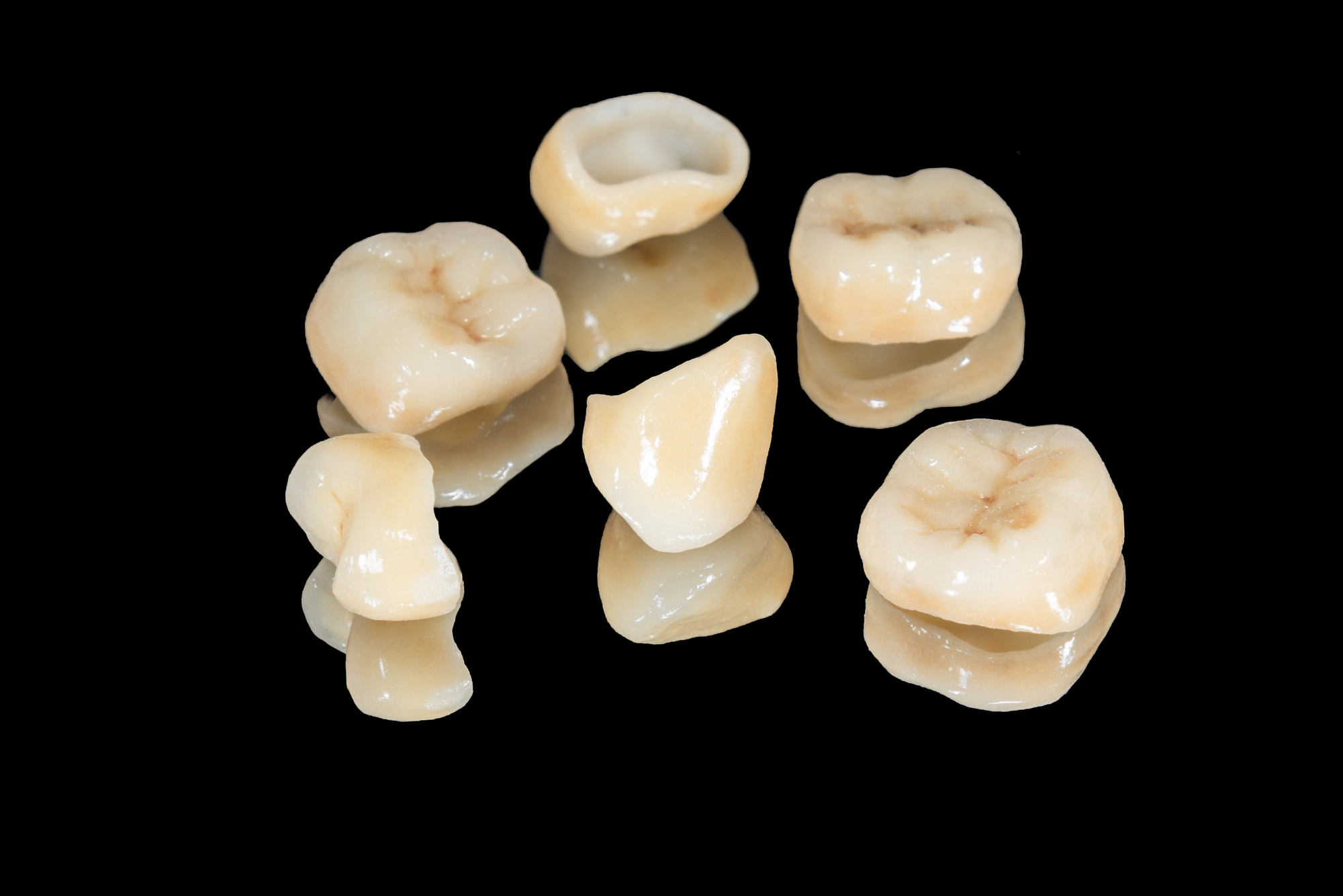 Seis coronas dentales de distintas formas y tamaños sobre una superficie negra reflectante.