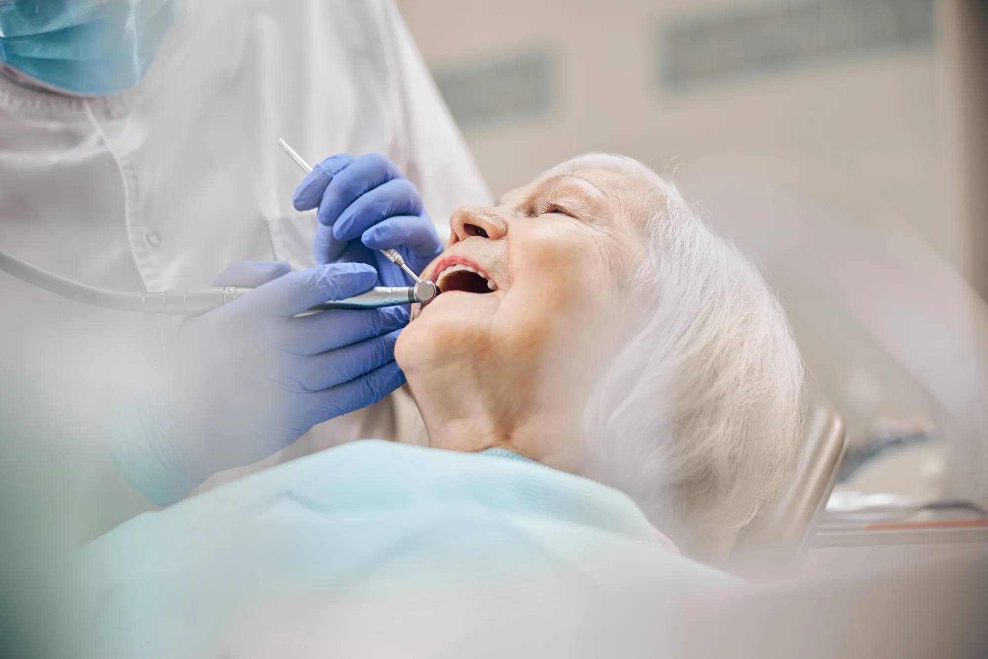 Dentist wearing gloves and a mask treating an elderly patient's teeth in a dental clinic.