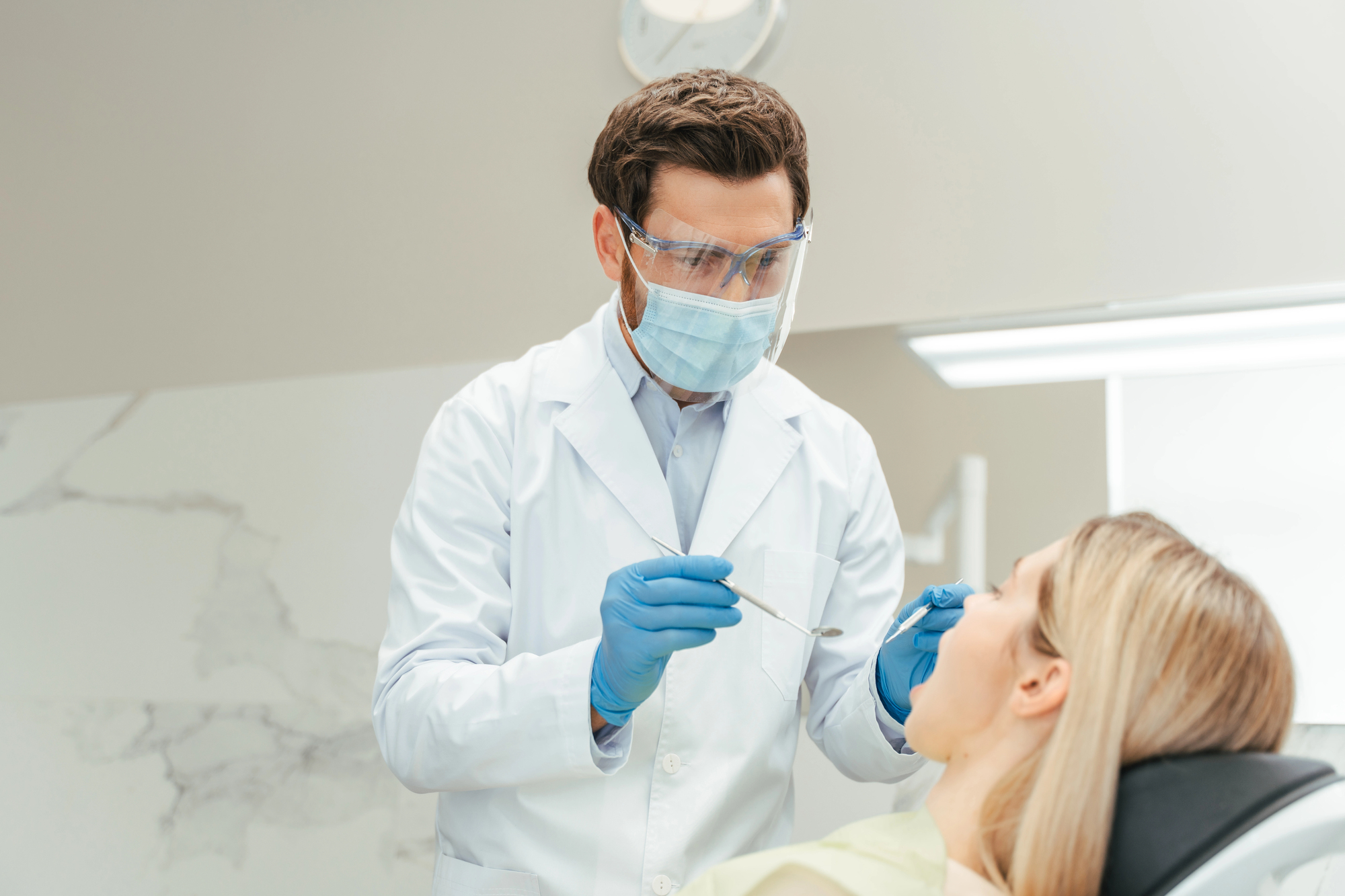 Dentist wearing protective mask, gloves, and face shield examining a female patient's mouth in a dental clinic.