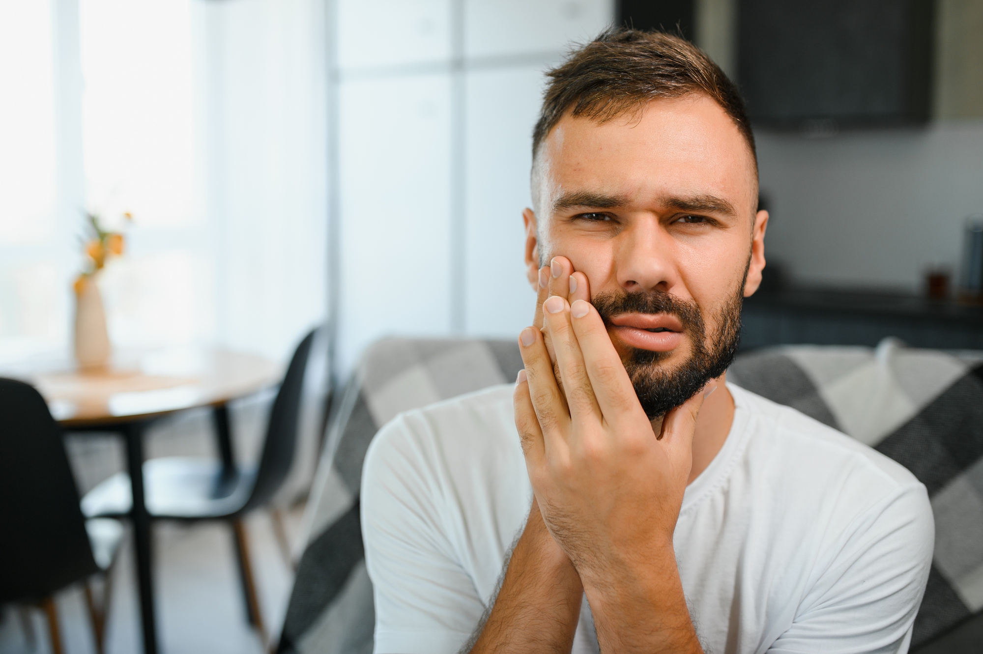 Man in white shirt holding his cheek with a painful expression, indicating toothache.