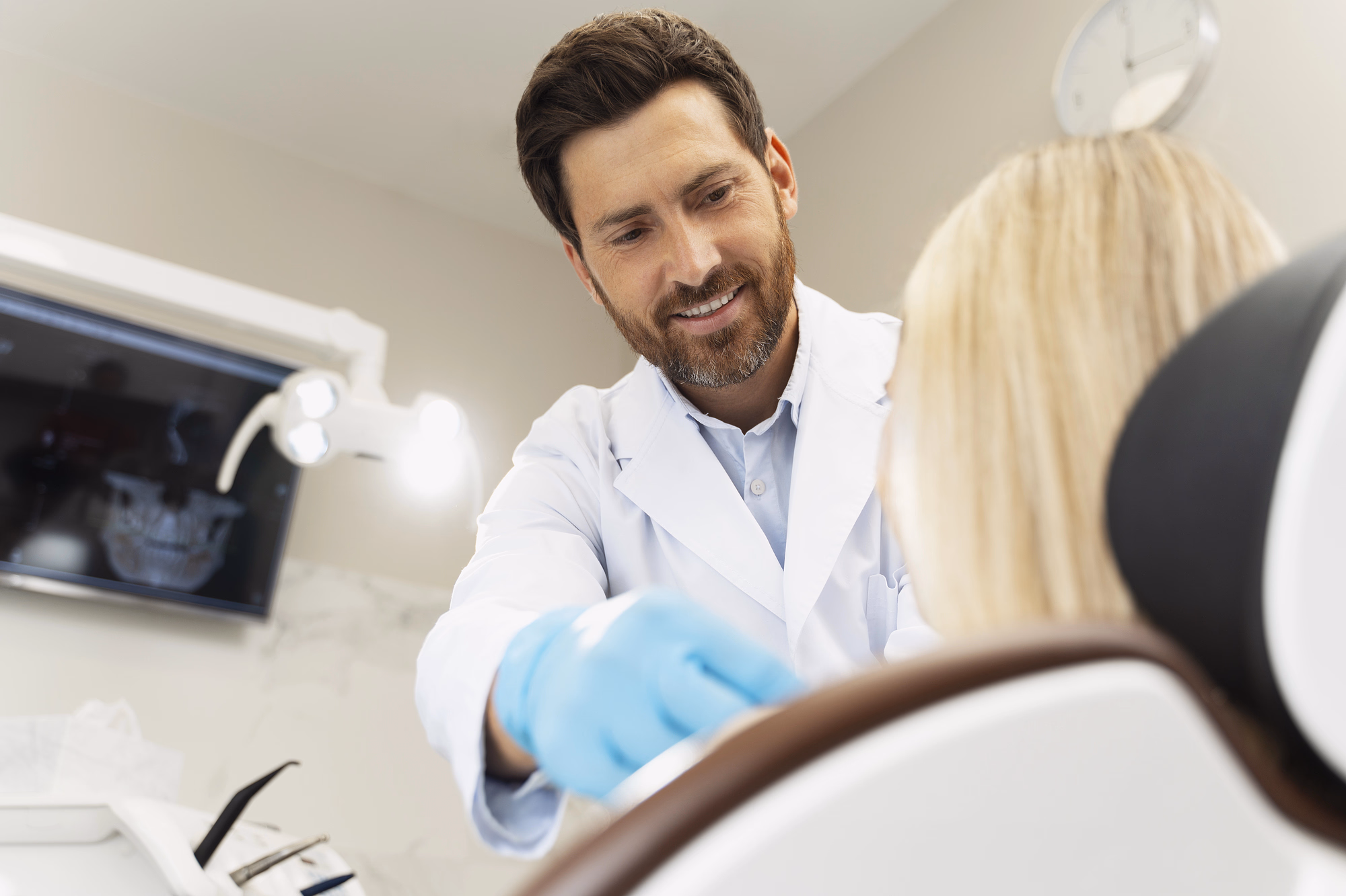 Male dentist in a white coat and blue gloves smiling and examining a female patient sitting in a dental chair.