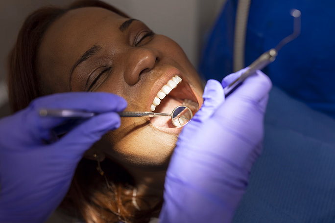 Dentist wearing purple gloves examining an open mouth of a smiling woman with a dental mirror and probe.