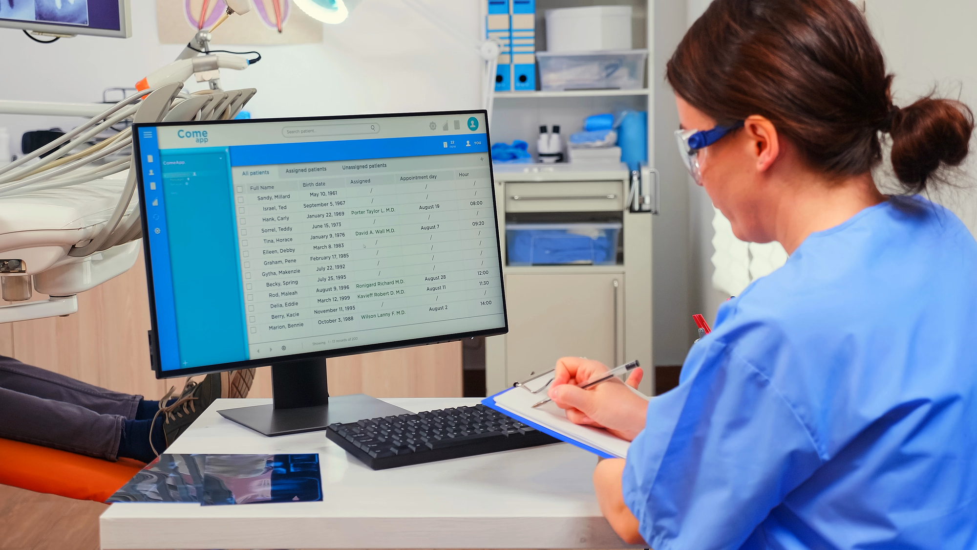 Healthcare professional in blue scrubs and protective glasses taking notes on a clipboard while reviewing patient appointment data on a computer screen.