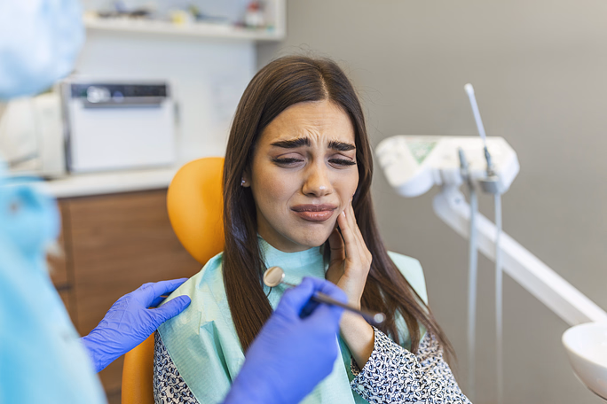 Woman in a dental chair holding her cheek in pain while a dentist wearing blue gloves comforts her.