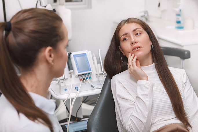 Mujer de cabello castaño largo sentada en una silla dental, sosteniéndose la mejilla con molestia, hablando con una dentista con bata blanca.