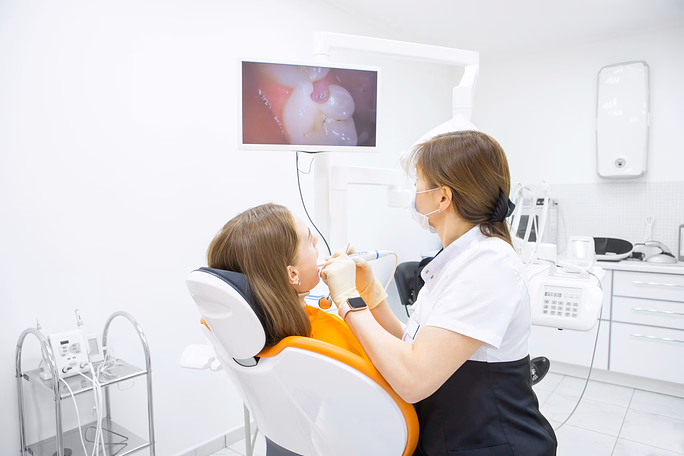 Dentist wearing a mask performing a dental procedure on a patient seated in an orange dental chair, with a close-up tooth image displayed on a monitor in a bright dental office.