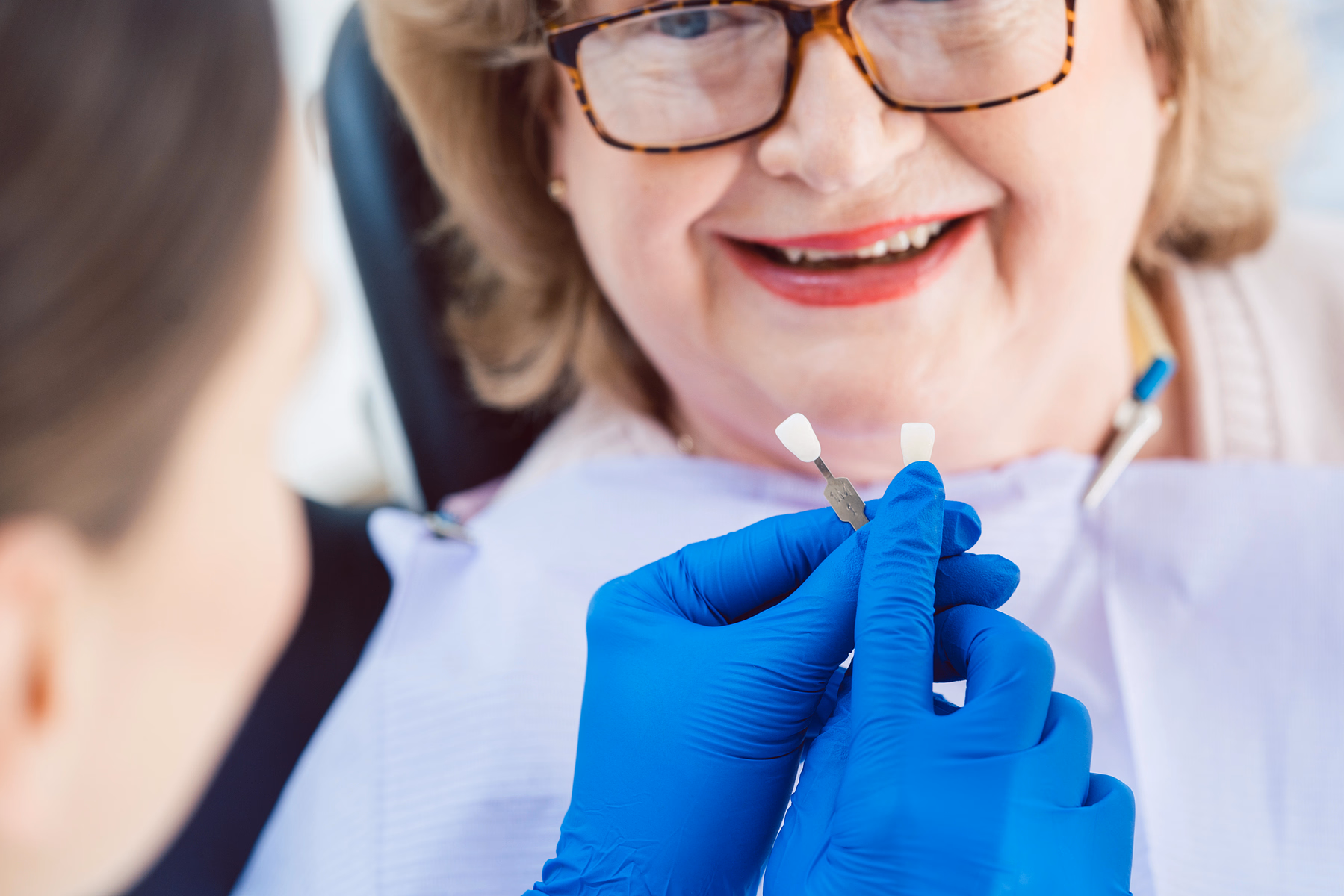 Dental professional with blue gloves holding two dental shade guides near a senior woman smiling in the dentist chair.