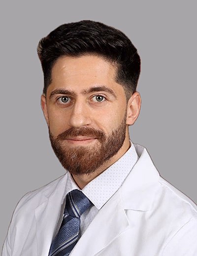 Retrato de un hombre con cabello y barba oscuros vistiendo una bata blanca de laboratorio, camisa de vestir y corbata azul a rayas sobre un fondo gris.