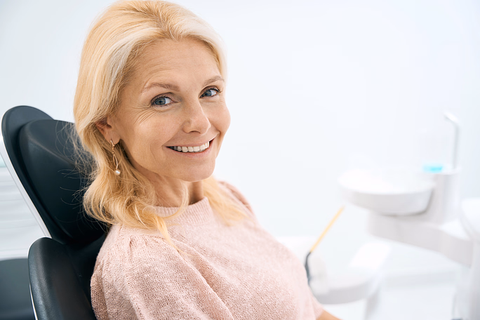 Mujer sonriente de cabello rubio sentada en una silla dental en una oficina dental luminosa.