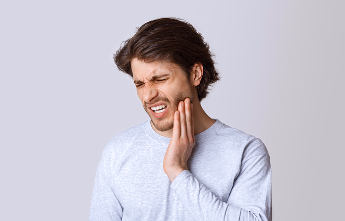 Young man in a light gray shirt holding his jaw with a pained expression, indicating toothache.