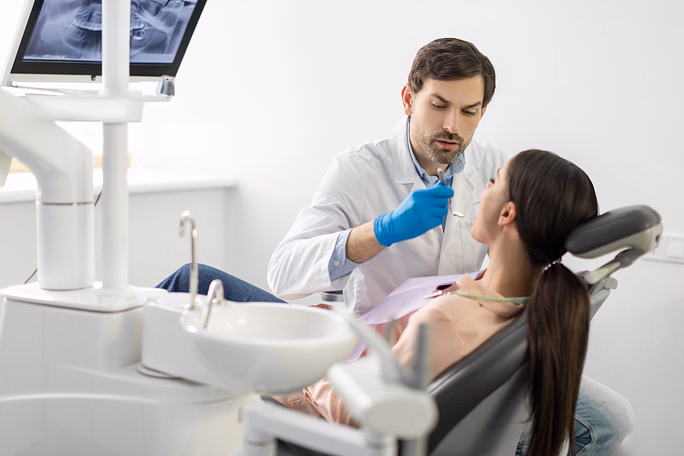 Dentist in white coat and blue gloves examining a female patient's mouth in a dental clinic.
