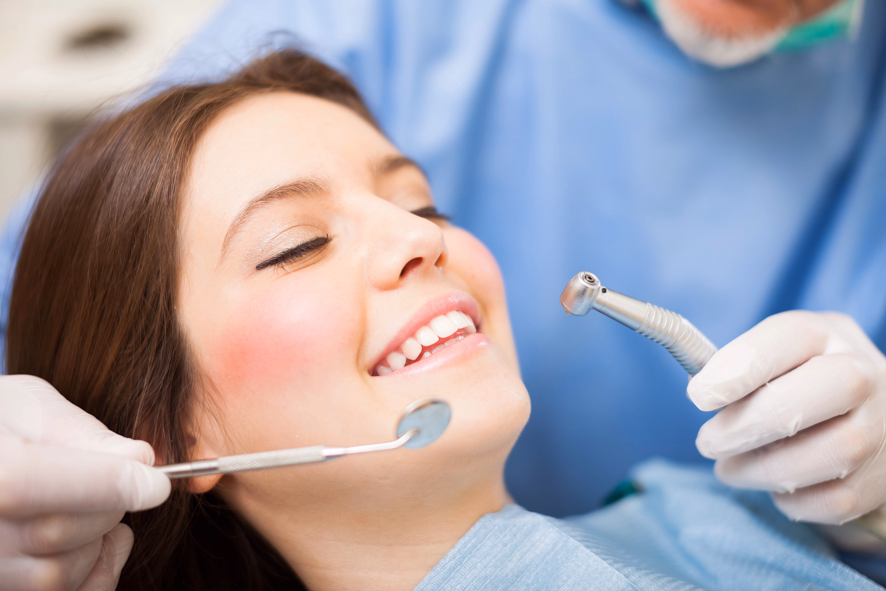 Smiling woman receiving dental checkup with dental mirror and drill held by dentist wearing gloves.