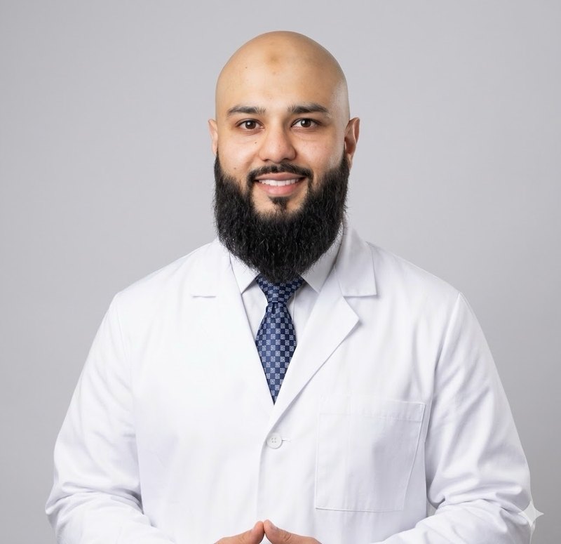 El Dr. Shehar Awan, dentista en American Urgent Dental, sonriendo con una bata blanca de laboratorio y corbata azul.