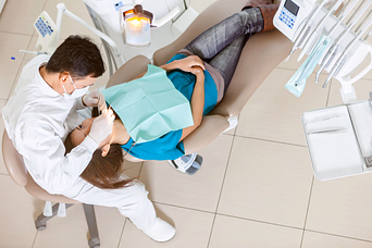Dentist in white coat examining patient's teeth while patient reclines in dental chair with a protective bib.