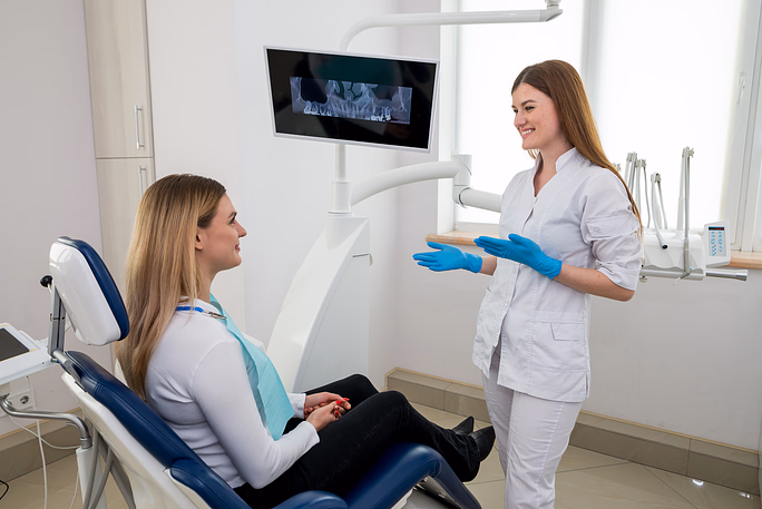 Dentista sonriente con guantes habla con una paciente sentada en una silla dental, con una radiografía dental mostrada en una pantalla.